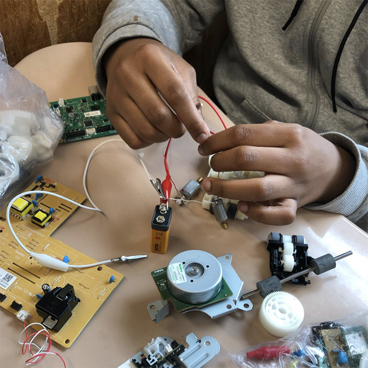 Closeup of electronic handling; alligator clips are being attached to a battery surrounded by deconstructed electronic parts. 